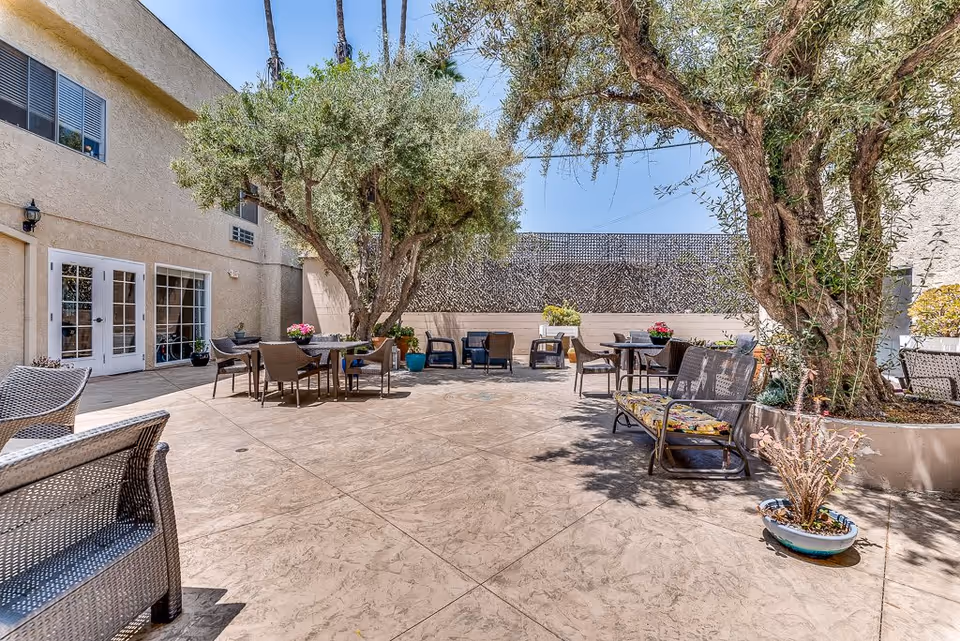 Outdoor patio area at Astoria Park Senior Living featuring several seating arrangements with tables and chairs, large trees providing shade, potted plants, and a textured privacy wall under a clear blue sky.