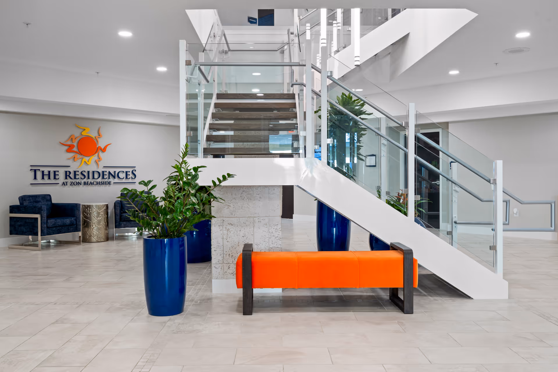 Modern interior lobby area of The Residences at Zon Beachside featuring a staircase with glass railings, an orange bench, blue planters with green plants, and two blue armchairs with a small metallic side table. The facility's logo is displayed on the wall.