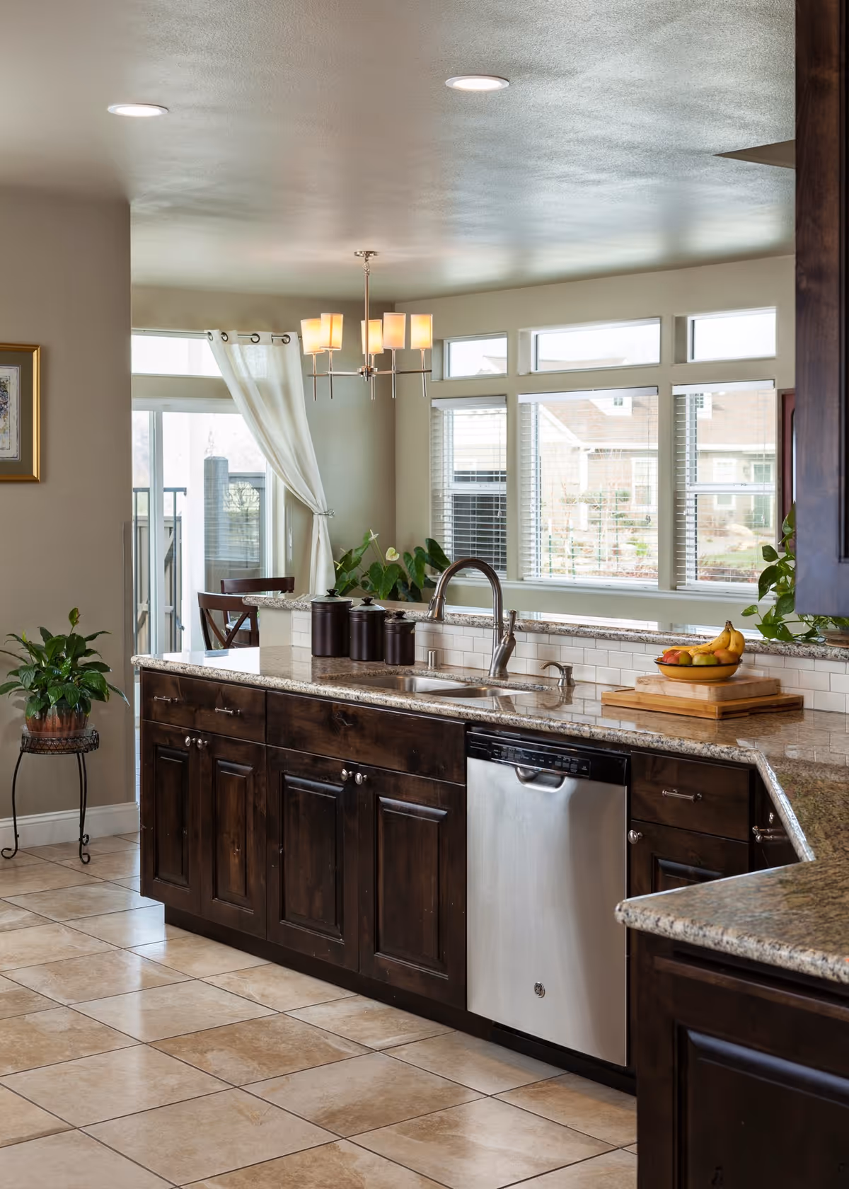 Bright kitchen with dark wooden cabinets, granite countertops, a stainless steel dishwasher, and a double sink with a modern faucet. There is a bowl of fruit on a wooden cutting board, three black canisters, and a plant on the counter. Large windows with white blinds and a glass door with white curtains let in natural light. Beige tiled floor and a chandelier with five lights hang from the ceiling.