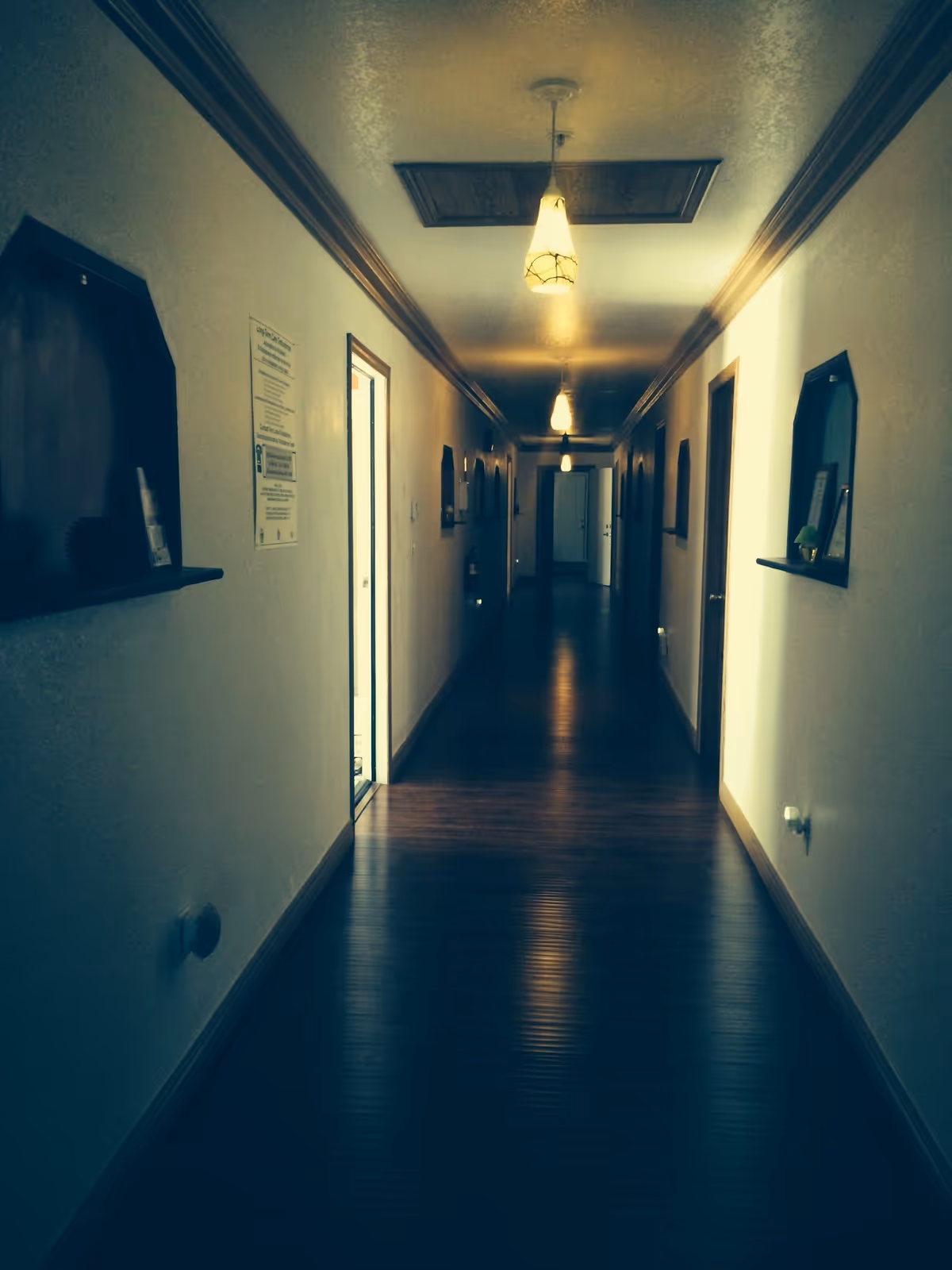 A long, dimly lit hallway with wooden flooring and white walls. The ceiling has recessed panels and hanging light fixtures. There are several doorways and small wall niches along the corridor.