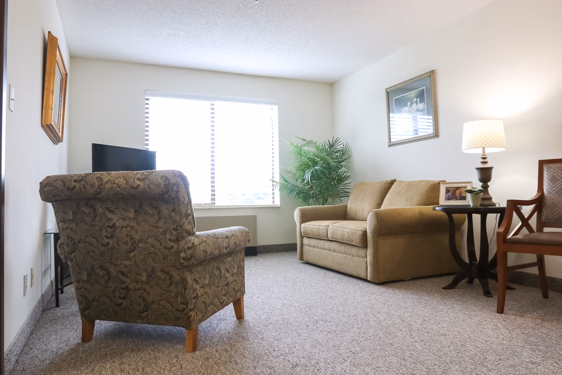 A bright living room with an upholstered armchair, loveseat, side table with a lamp and framed photo, and a large window.