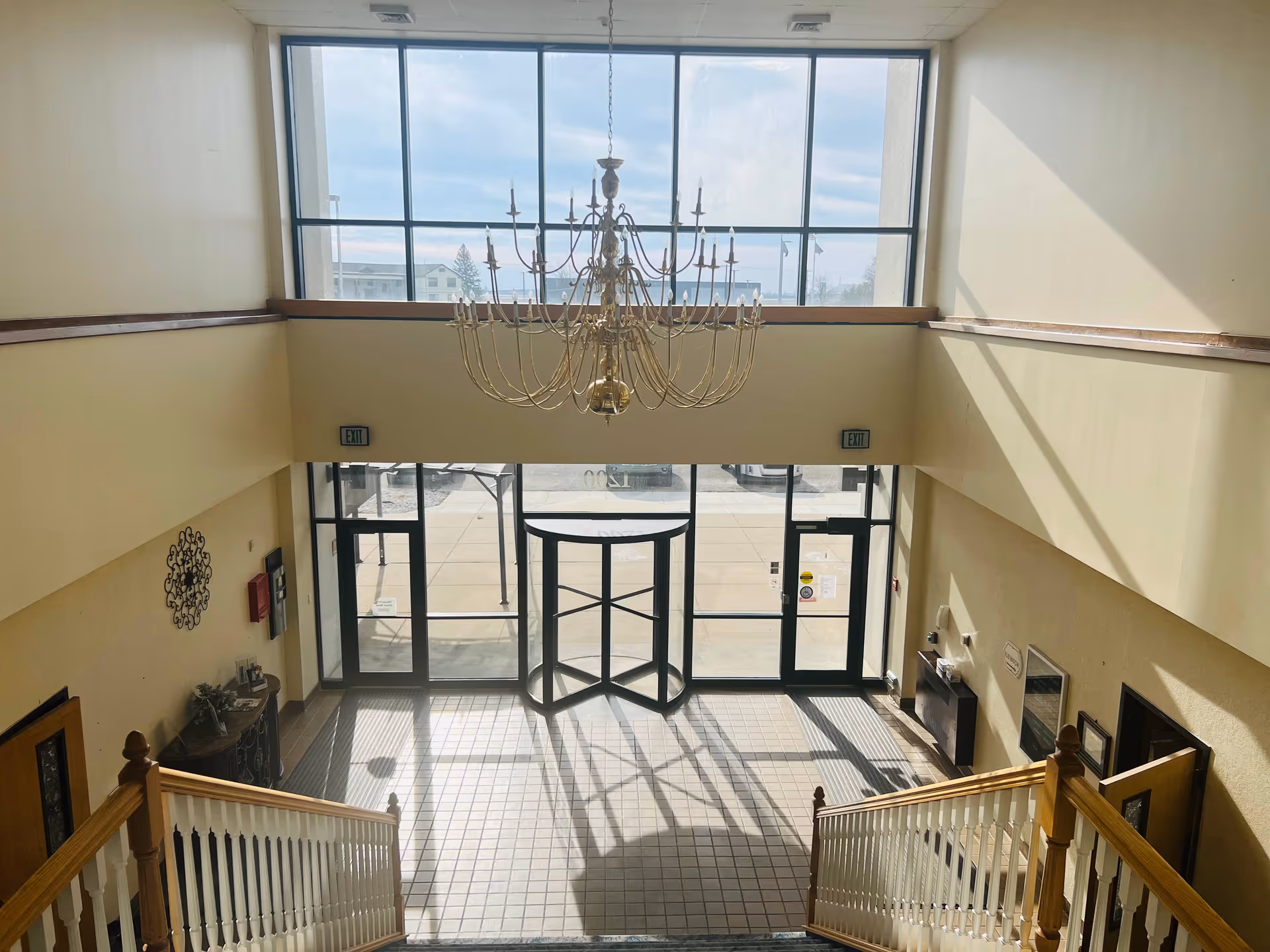 View from the top of a staircase looking down into a spacious lobby area with large windows and a glass revolving door. A large chandelier hangs from the ceiling, and sunlight casts shadows on the tiled floor. There are two exit doors on either side of the revolving door and some decorative elements on the walls.