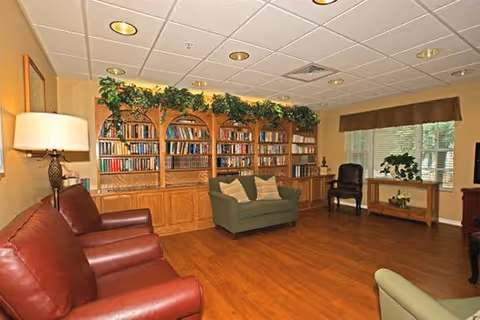 A cozy communal living room with built-in bookshelves, seating (armchairs and a loveseat), plants, and wood flooring.