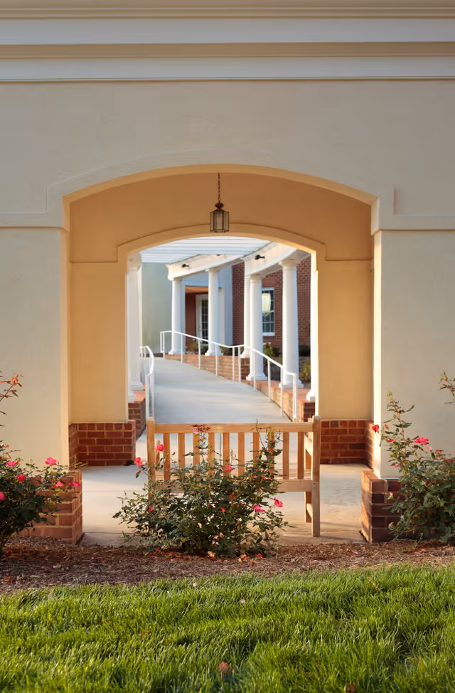 A wooden bench surrounded by rose bushes sits under a beige archway with a hanging lantern. Beyond the archway is a covered walkway with white columns and a ramp leading to a brick building.