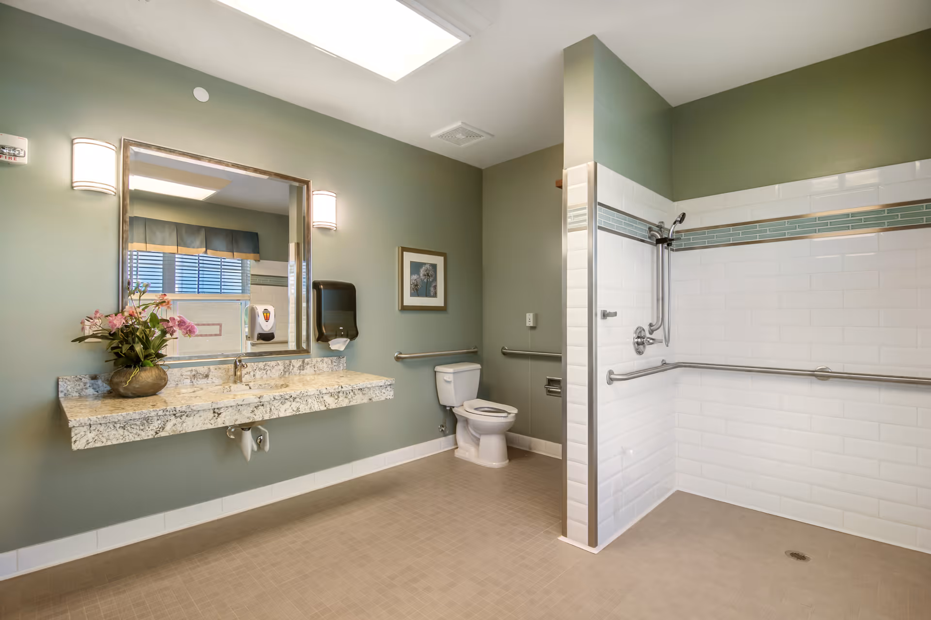 A spacious bathroom with green walls and beige tiled floor. It features a large granite countertop with a sink, a mirror with two wall-mounted lights, a toilet with grab bars, and a walk-in shower with white subway tiles and a horizontal decorative tile strip. There is a flower arrangement on the countertop and a framed picture on the wall.