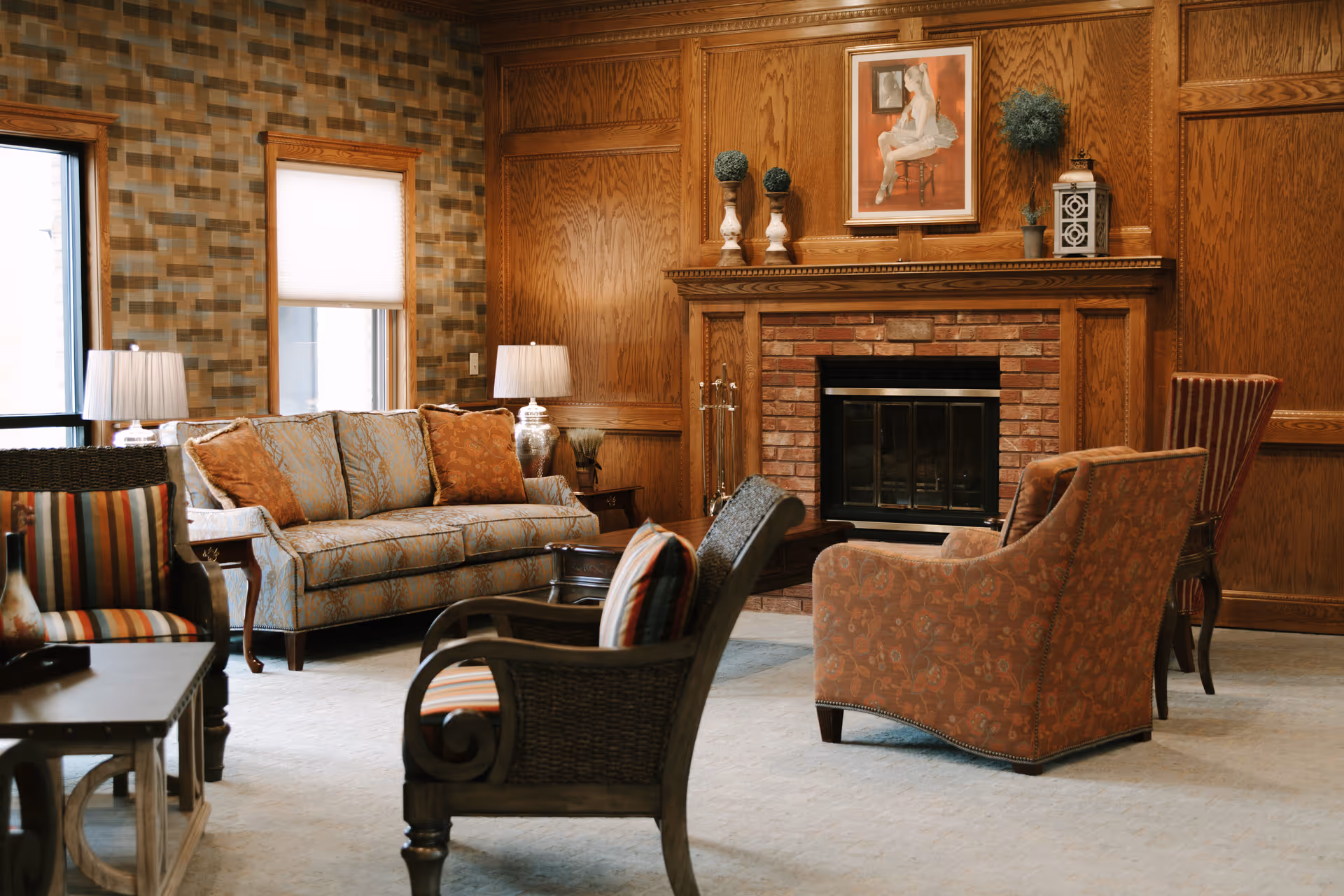 A cozy living room with a brick fireplace surrounded by wooden paneling. The room features a patterned sofa with orange cushions, two armchairs with striped upholstery, a cushioned armchair with floral patterns, a wooden coffee table, two table lamps, and decorative items on the fireplace mantel including vases, a lantern, and a framed painting.