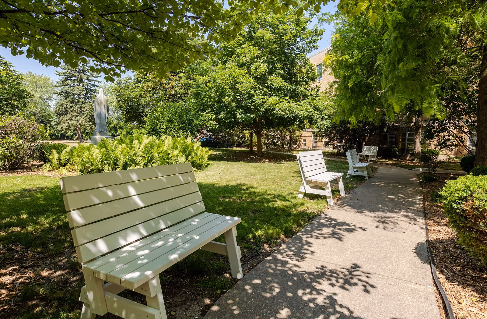 A peaceful outdoor garden area with a paved walkway lined with white wooden benches. The garden is lush with green trees, bushes, and ferns, and a statue is visible in the background. Sunlight filters through the leaves, creating dappled shadows on the ground.