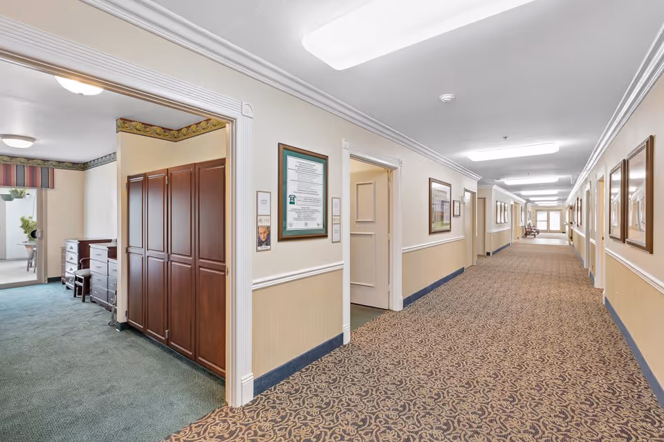 A long, well-lit hallway in a senior living facility with patterned carpet and beige walls adorned with framed pictures. On the left side, there is an open doorway leading to a room with green carpet, wooden cabinets, and a window with a striped valance. The hallway extends into the distance with multiple doors and a bench near the far end.