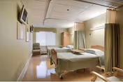 Two hospital-style beds in a bright skilled nursing patient room with privacy curtains, chairs, and a wall-mounted TV.
