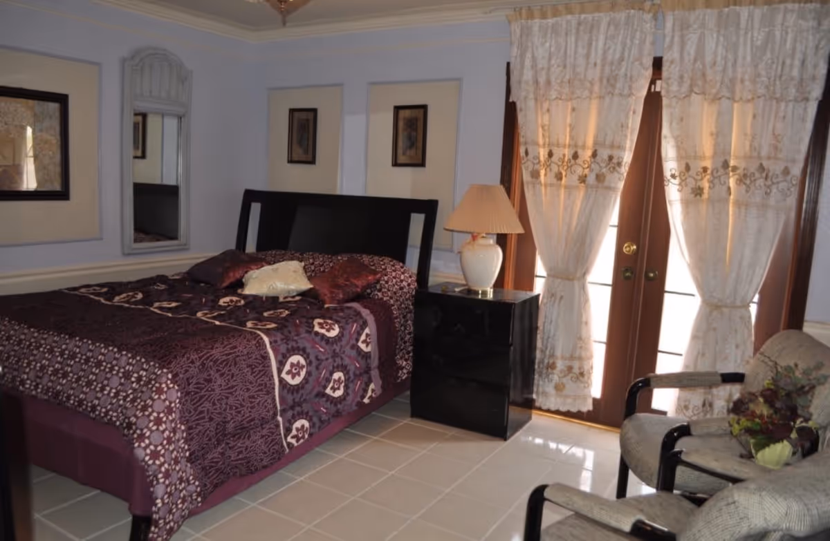 A bedroom with a large bed covered in a purple patterned bedspread and several pillows. There is a black nightstand with a white lamp next to the bed. Two cushioned chairs with black armrests are positioned near a small table with a plant. The room has light-colored tiled flooring, light blue walls with framed artwork, a mirror, and a set of French doors with sheer, embroidered curtains.