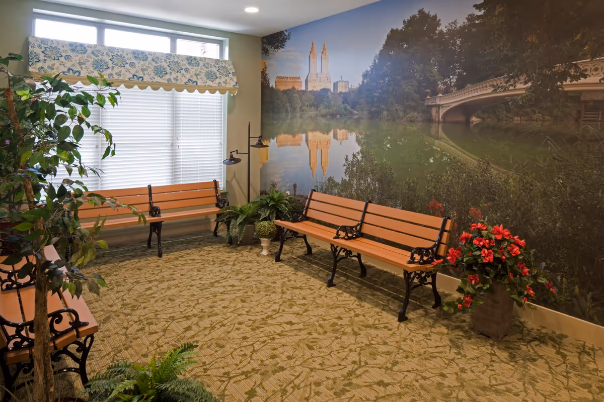 Indoor seating area with wooden benches featuring black metal armrests and legs, surrounded by various potted plants. A large wall mural depicts a serene lake scene with trees, a bridge, and tall buildings in the background. A window with white blinds and a floral valance allows natural light into the room.