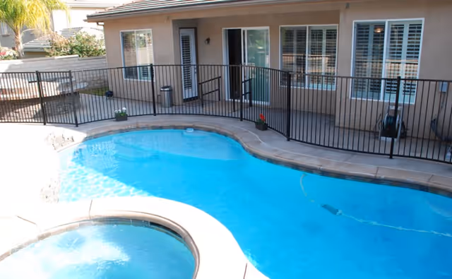 Outdoor swimming pool and hot tub area enclosed by a black metal safety fence, adjacent to a beige building with multiple windows and a sliding glass door.