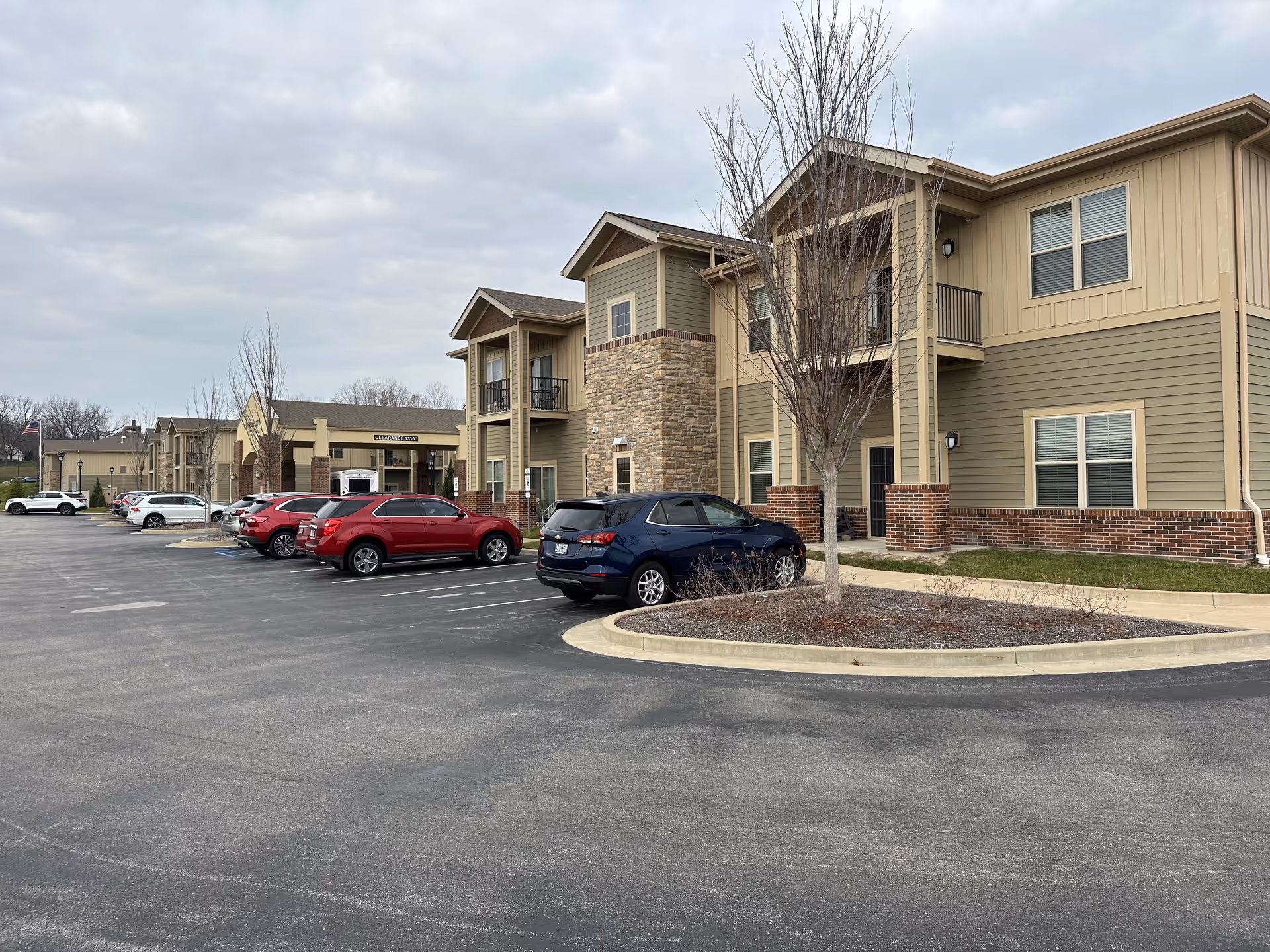 Exterior view of Boulevard Senior Living Of St Charles showing a two-story building with beige and brown siding, brick accents, and several windows. There is a parking lot in front with multiple parked cars and a few leafless trees in landscaped areas. The sky is overcast.