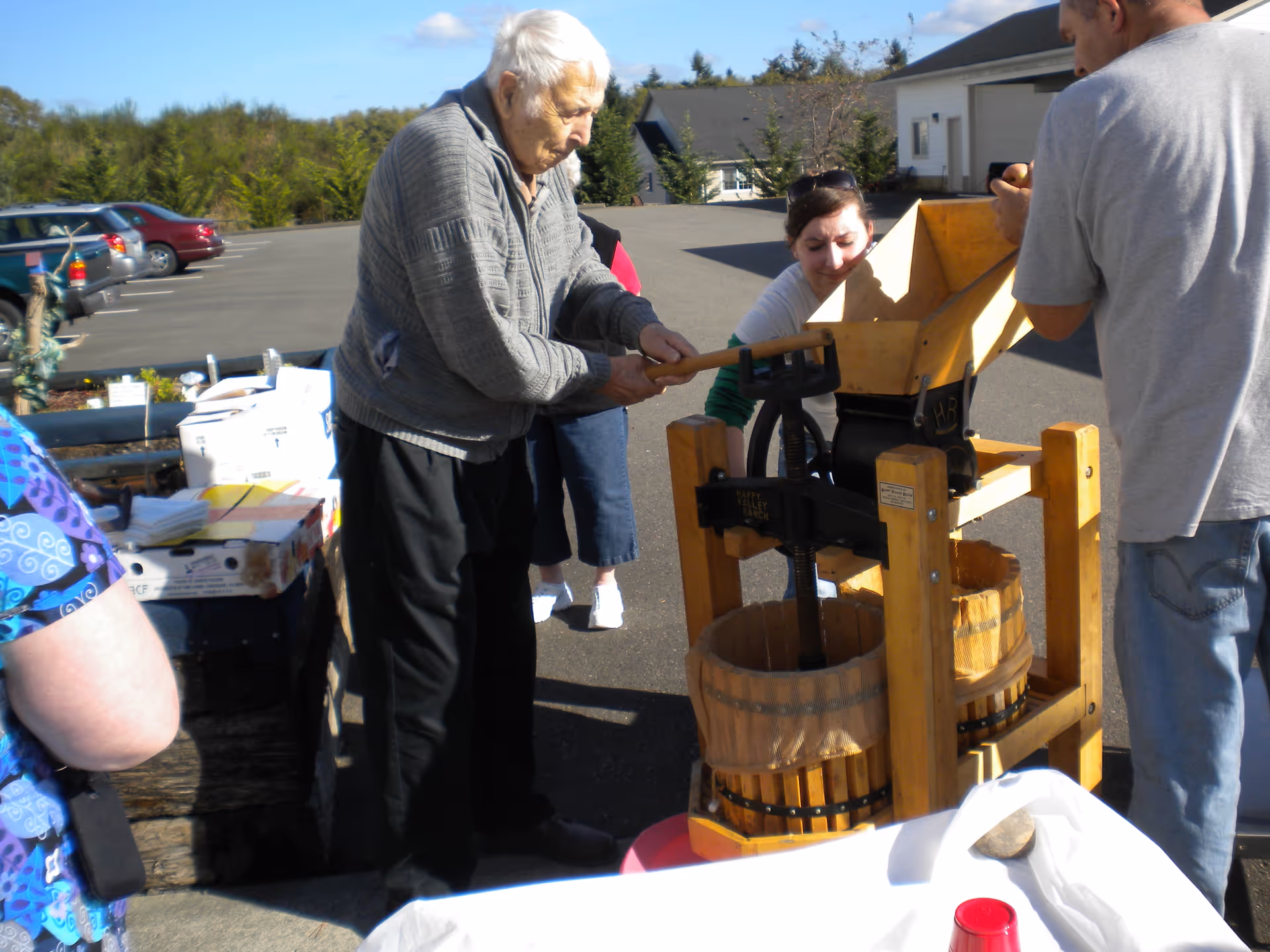 An elderly man is operating a wooden fruit press outdoors with assistance from two other people. There are cars parked in the background and a building visible under a clear blue sky.