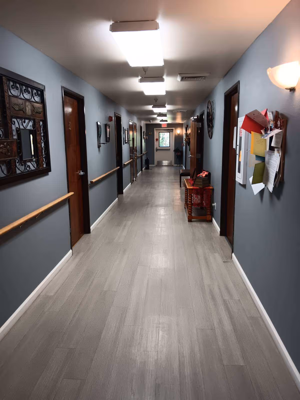Long, well-lit interior hallway with gray walls, wooden doors, handrails and a bulletin board in a senior living facility.