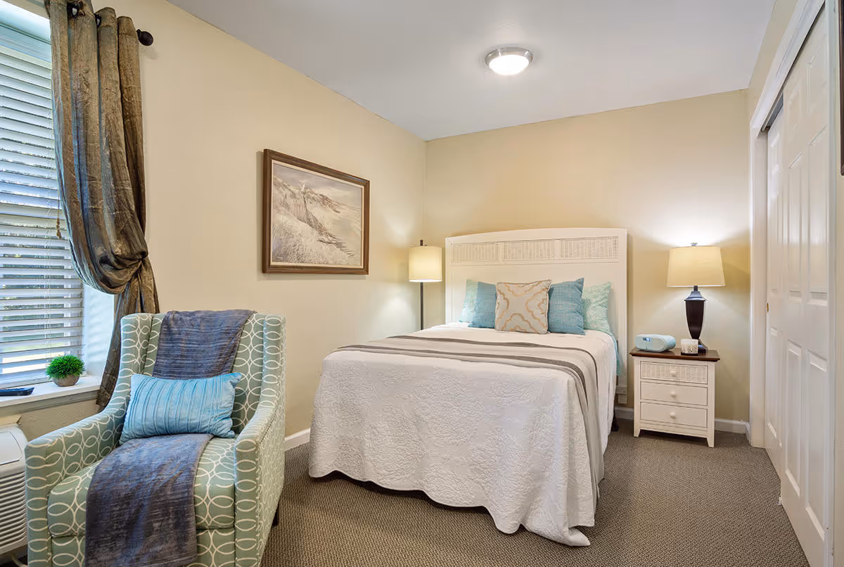 A cozy bedroom with a white bed featuring multiple pillows and a white quilt. To the right of the bed is a nightstand with a lamp, a clock, and a small box. On the left side of the room, there is a patterned armchair with a blue throw and pillow next to a window with blinds and dark curtains. A framed landscape painting hangs on the wall above the bed. The room has beige walls and carpeted flooring.