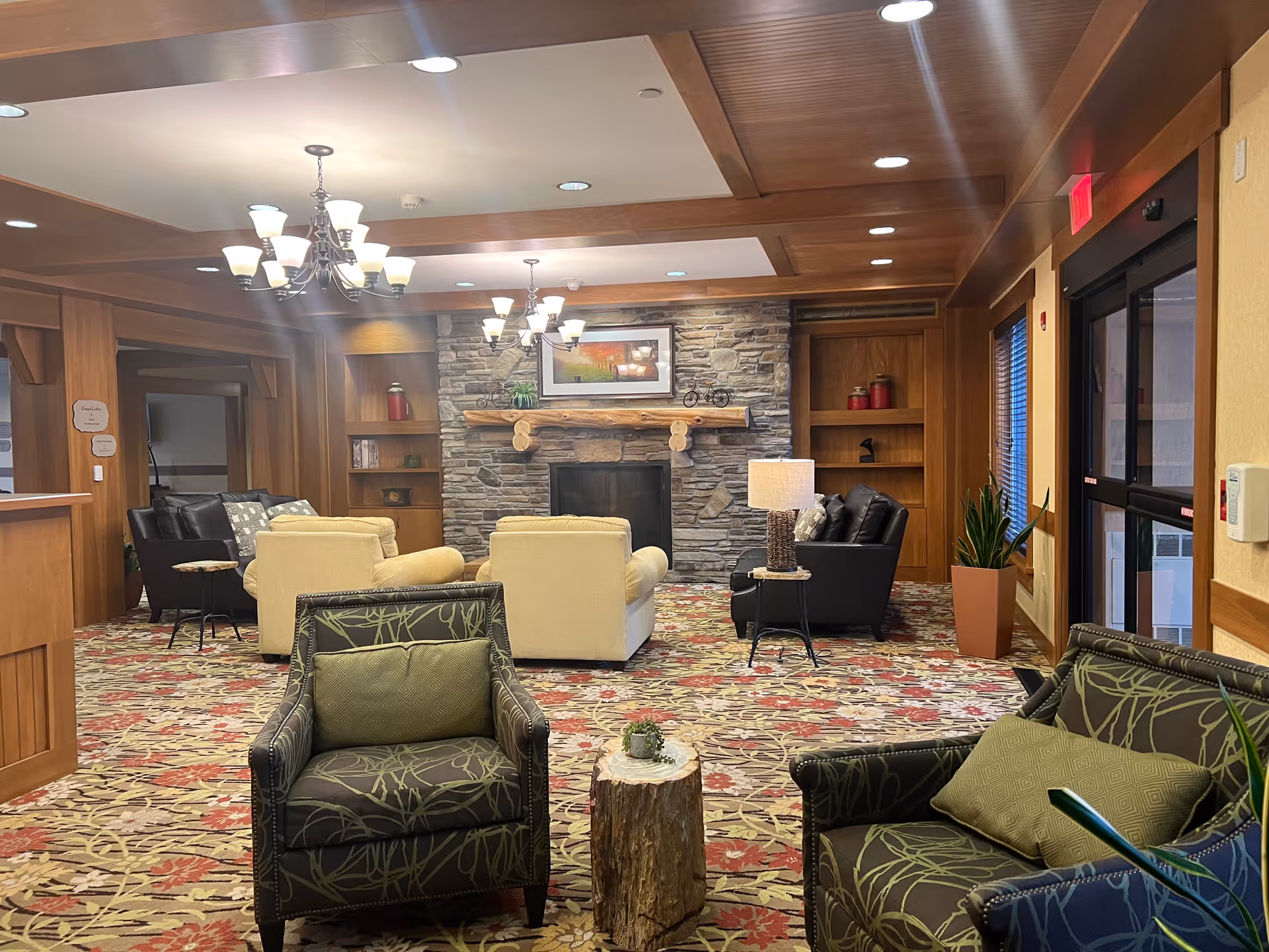 A cozy lobby-style common room with upholstered chairs and sofas arranged around a stone fireplace under chandelier lighting.