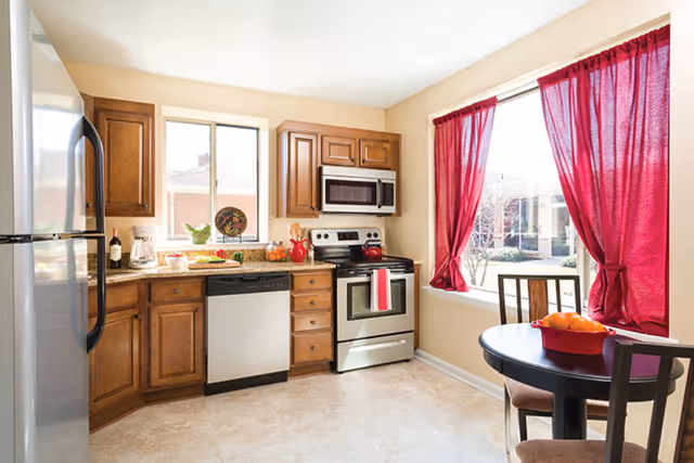 Sunlit kitchen with wooden cabinets, stainless steel appliances, a small round dining table, and red curtains at the window.