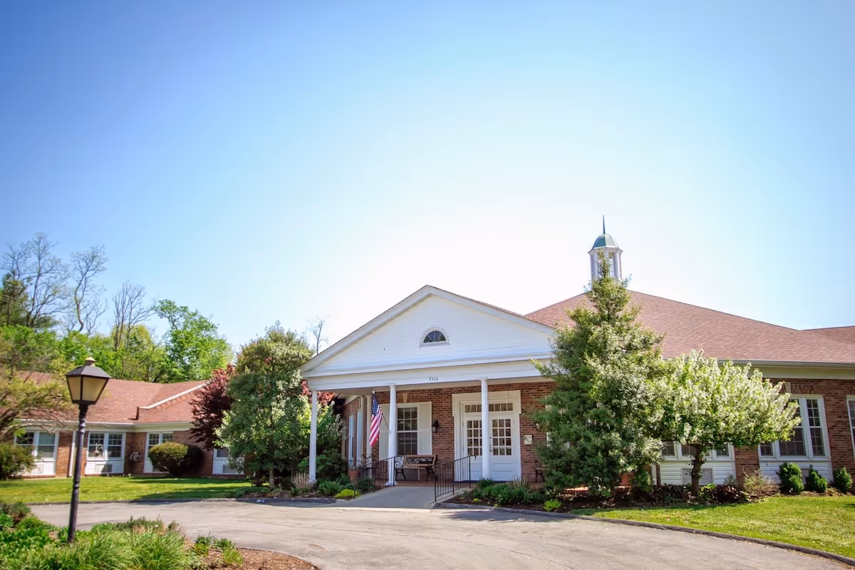 Exterior view of Mayfair Manor, a single-story brick building with a white entrance porch, surrounded by green trees and shrubs under a clear blue sky.