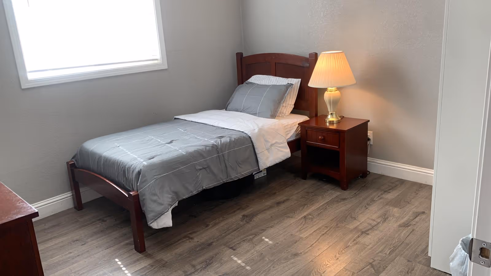 A small furnished bedroom in a senior care facility with a single bed, nightstand, and a lamp.