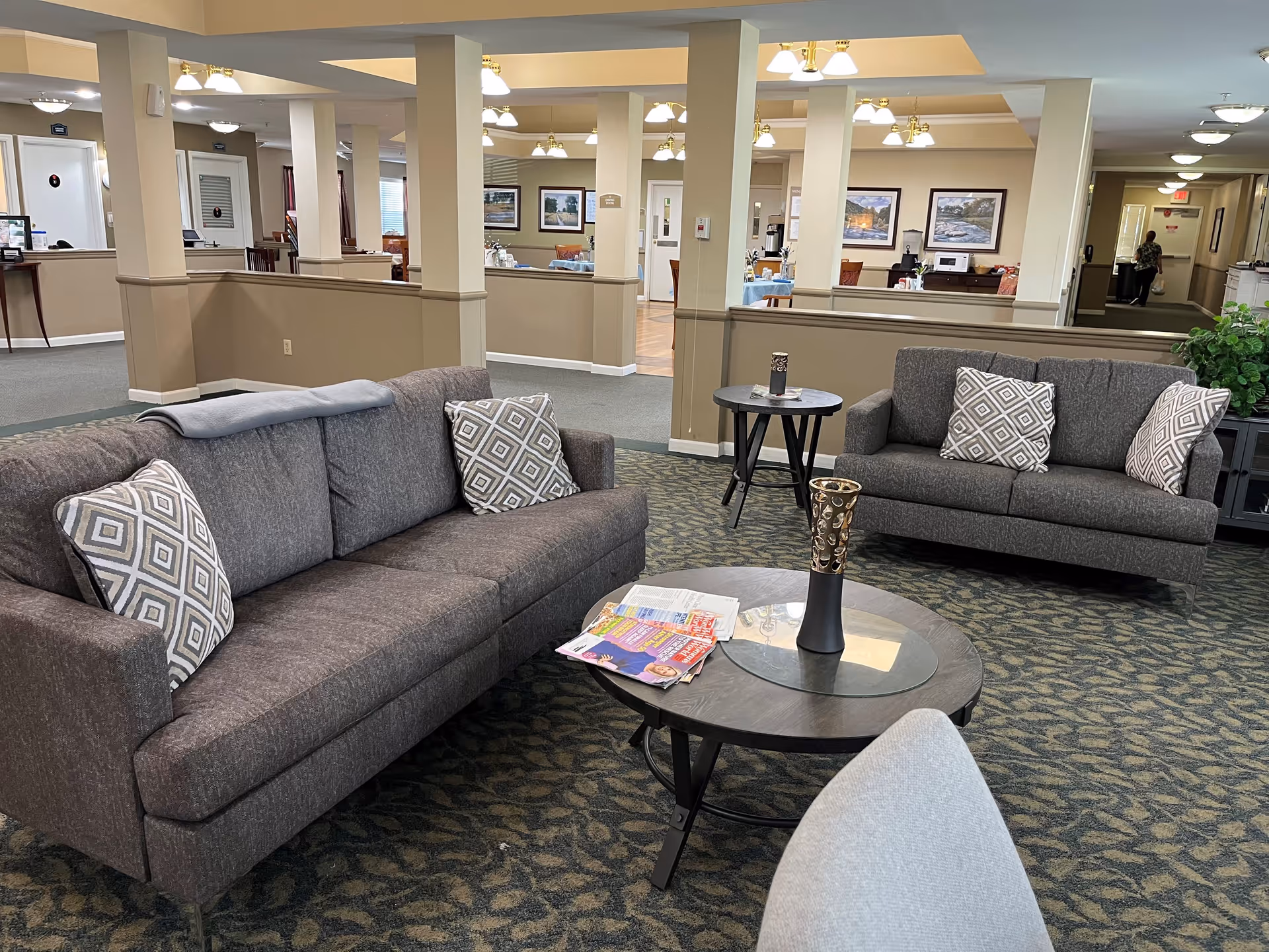 A cozy living room area in a senior living facility with two gray sofas adorned with patterned throw pillows, a round coffee table with a decorative vase and magazines, and a small side table. The background shows an open interior space with beige walls, columns, framed pictures, and ceiling lights.