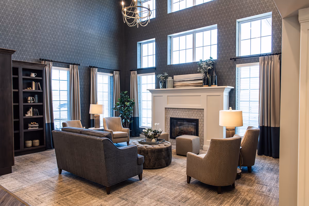 A cozy communal lounge with sofas and armchairs arranged around a fireplace beneath tall windows.