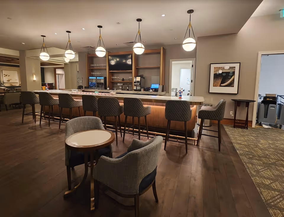 Modern communal bar and lounge area with a row of high stools at a counter, pendant lights, and small seating table in the foreground.