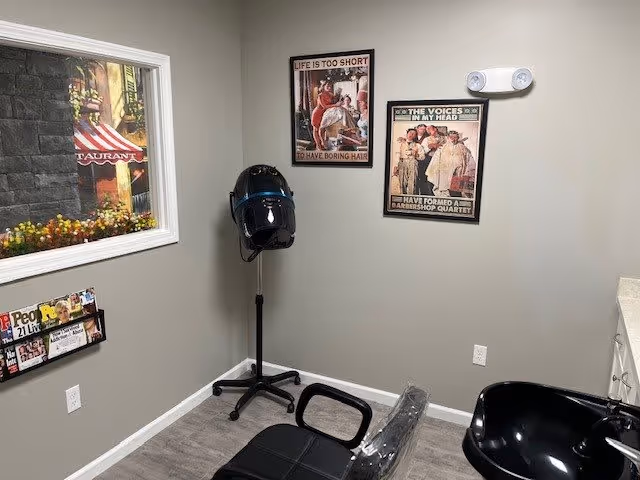Interior view of a small hair salon or barber area with a black salon chair, a black hair dryer hood on a stand, two humorous framed posters on the wall, a magazine rack with magazines, and a window showing an outdoor scene with flowers and a restaurant sign.