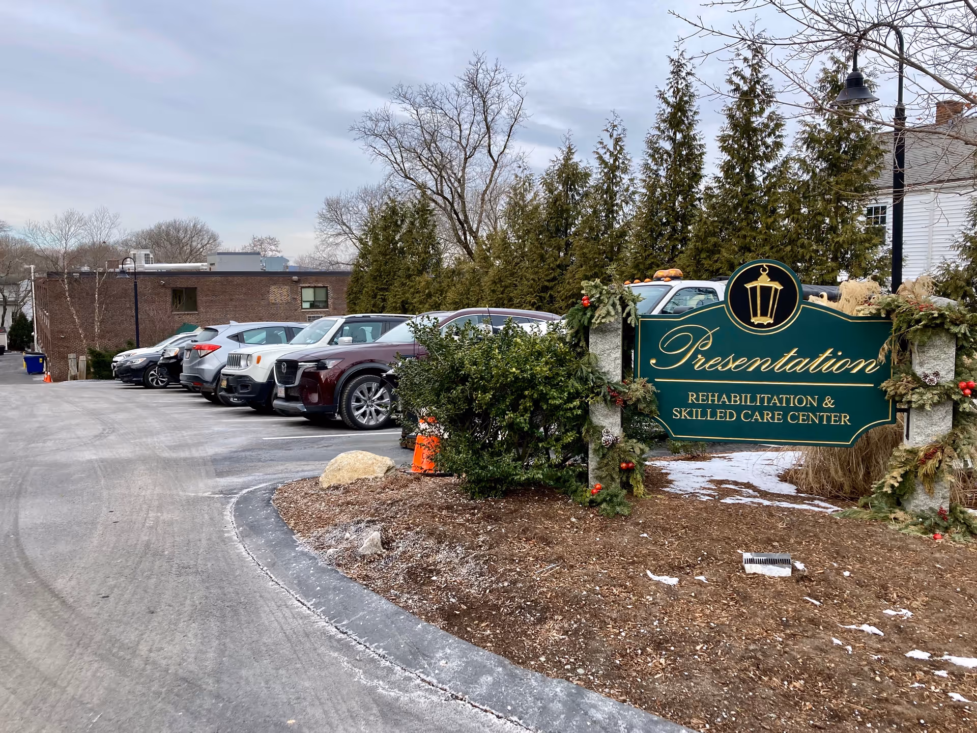 Parking lot with several parked cars next to a green and gold sign that reads 'Presentation Rehabilitation & Skilled Care Center' decorated with holiday greenery and ornaments, with trees and buildings in the background under a cloudy sky.
