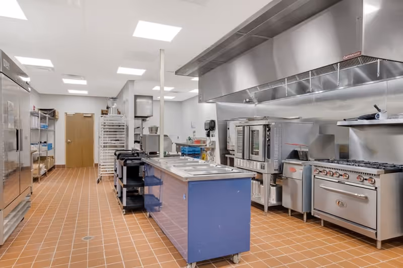 A clean, professional commercial kitchen with stainless steel appliances including ovens, stoves, and a large ventilation hood. There is a blue island counter with built-in food warmers in the center, and shelves with kitchen supplies along the walls. The floor is covered with reddish-brown tiles and the ceiling has multiple bright fluorescent lights.