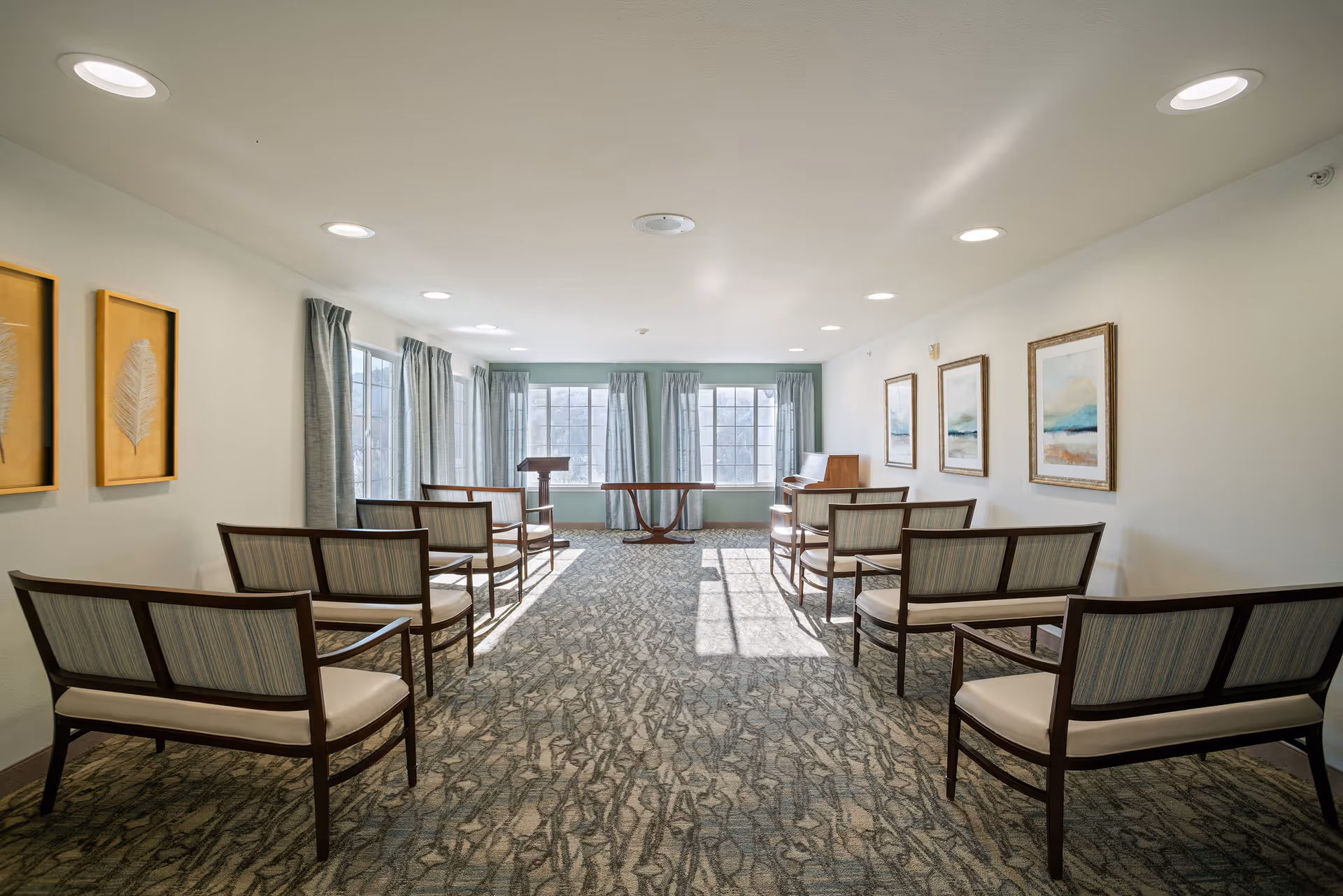 A bright room with large windows covered by light curtains, arranged with rows of wooden chairs with cushioned seats facing a small wooden podium and table at the front. The walls are decorated with framed artwork and the floor is carpeted with a patterned design.