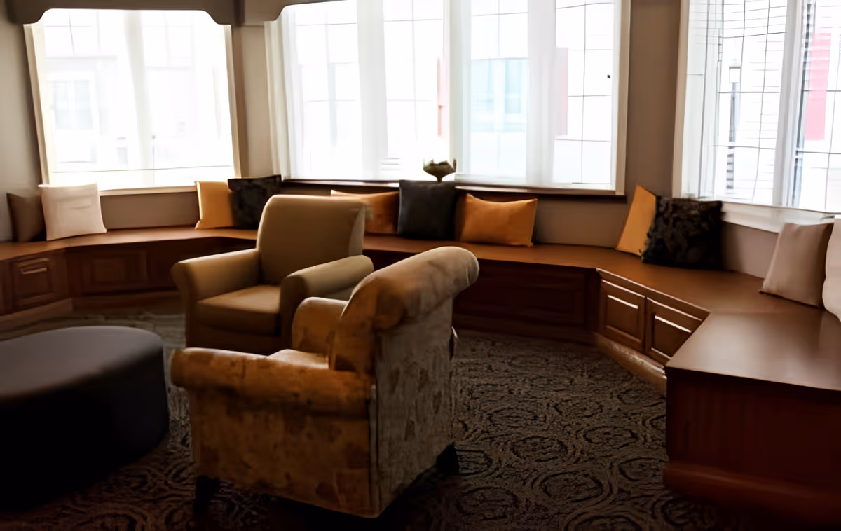 A cozy seating area with two armchairs and a large built-in wooden bench lined with cushions in front of tall windows with sheer white curtains, illuminated by natural light.