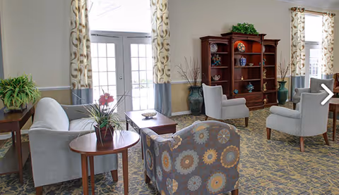 Sunny communal living room with upholstered sofas and chairs around coffee tables, a wooden display cabinet, and French doors with patterned curtains.