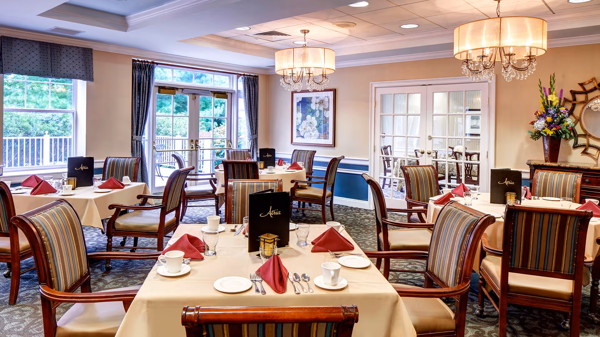 A well-lit dining room in a senior living facility with several tables set with white tablecloths, red folded napkins, cups, plates, and silverware. The chairs have wooden frames with striped upholstery. Large windows and glass doors allow natural light to enter, and there are two chandeliers hanging from the ceiling. A floral arrangement and framed artwork decorate the room.