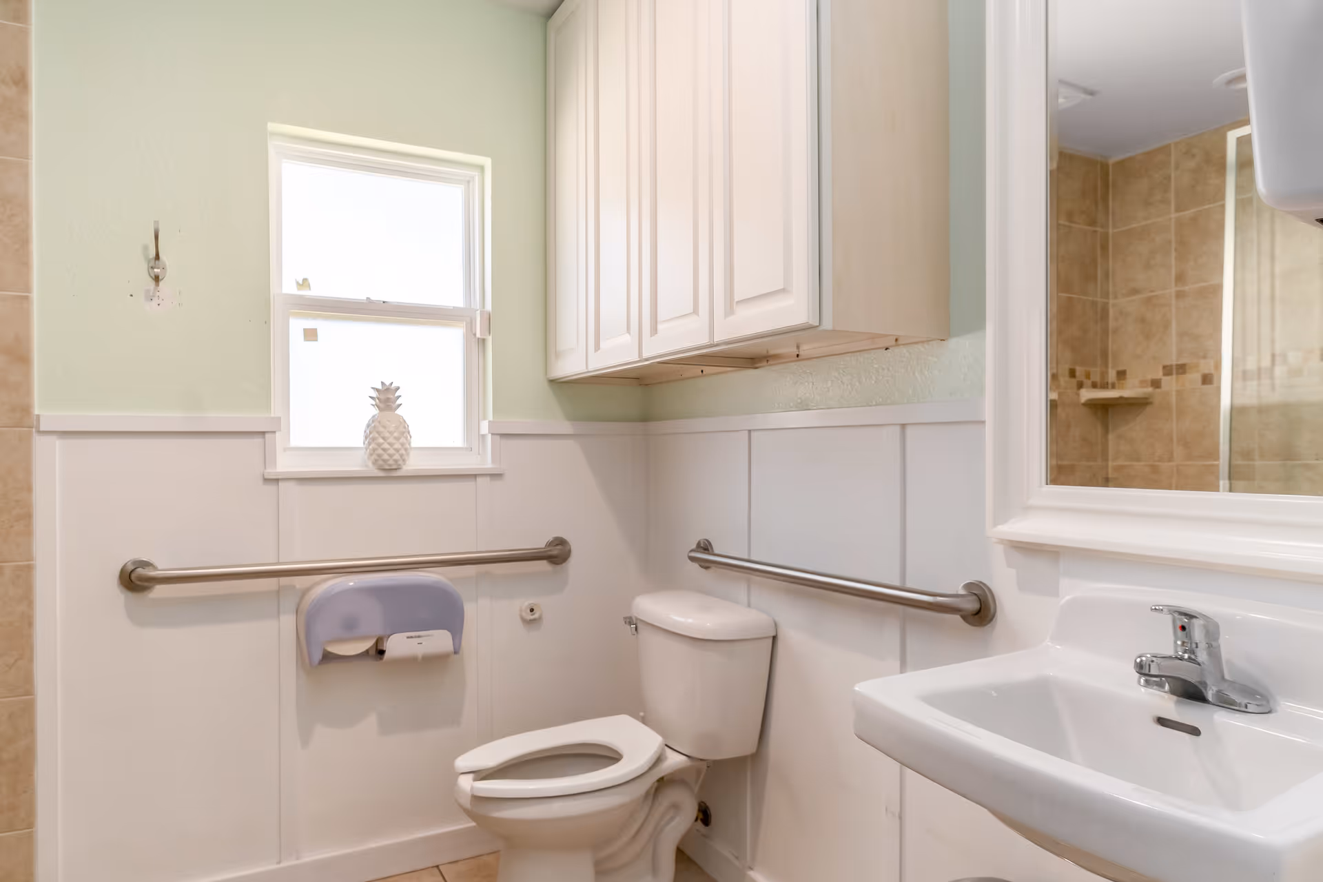 Bright accessible bathroom featuring a toilet, sink, stainless grab bars, wall cabinets and a window with a decorative pineapple.