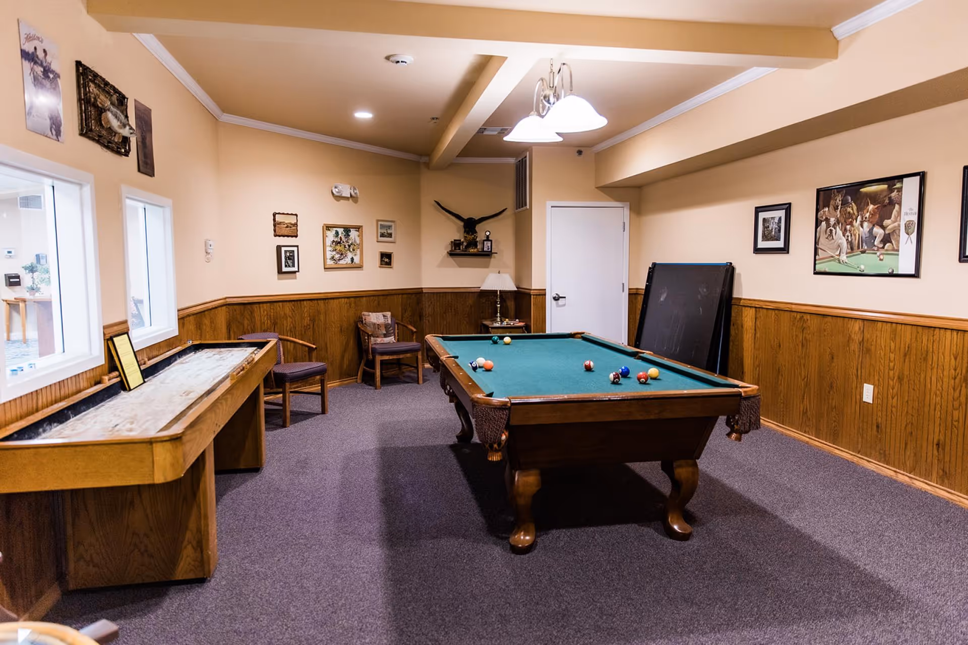 A recreation room with a pool table and a shuffleboard table. The room has beige walls with wood paneling on the lower half, carpeted floor, and several framed pictures on the walls. There are two chairs and a small table with a lamp in the corner. Ceiling lights illuminate the room.
