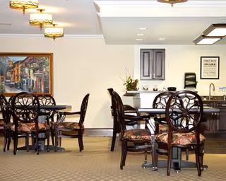 Dining room with dark wooden tables and chairs, patterned carpet, artwork on the wall and a small kitchenette/bar area under warm lighting.
