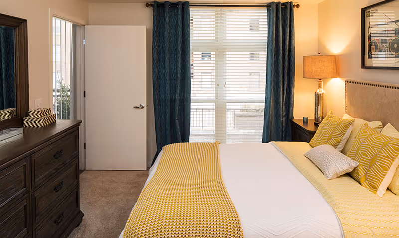 Well-lit bedroom with a made bed dressed in yellow and white bedding, a dresser and mirror on the left, a bedside table with lamp, and a window with teal curtains.
