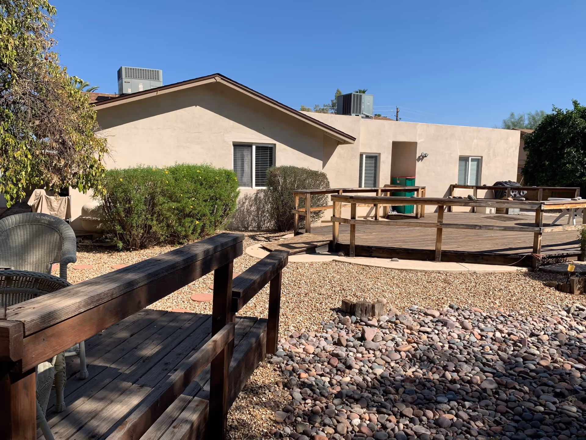 Outdoor area of a senior living facility with a wooden deck and ramp, surrounded by gravel and rocks. There are bushes and trees near a beige building with windows and air conditioning units on the roof under a clear blue sky.