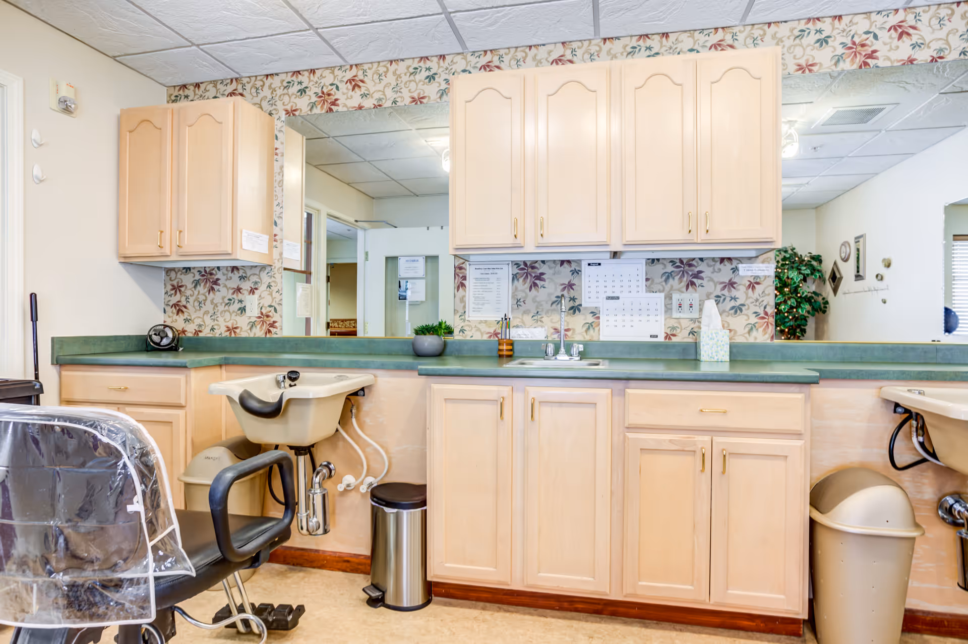 Interior view of a hair salon area within a senior living facility, featuring a salon chair with a plastic cover, a hair washing sink, light wood cabinets, a green countertop, floral wallpaper, and a large mirror reflecting part of the room.