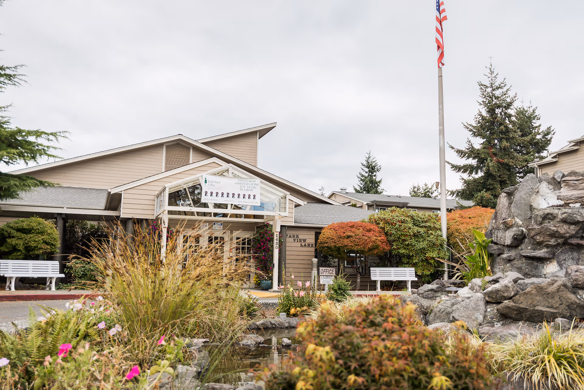 Front entrance of a senior living facility with landscaping, benches, a flagpole, and a pond-like water feature.