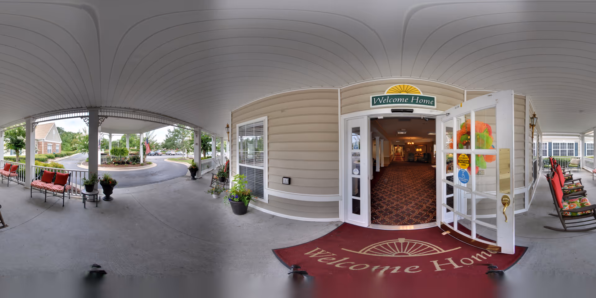 Covered front porch and entrance of a senior living facility with open double doors, a 'Welcome Home' mat and seating.