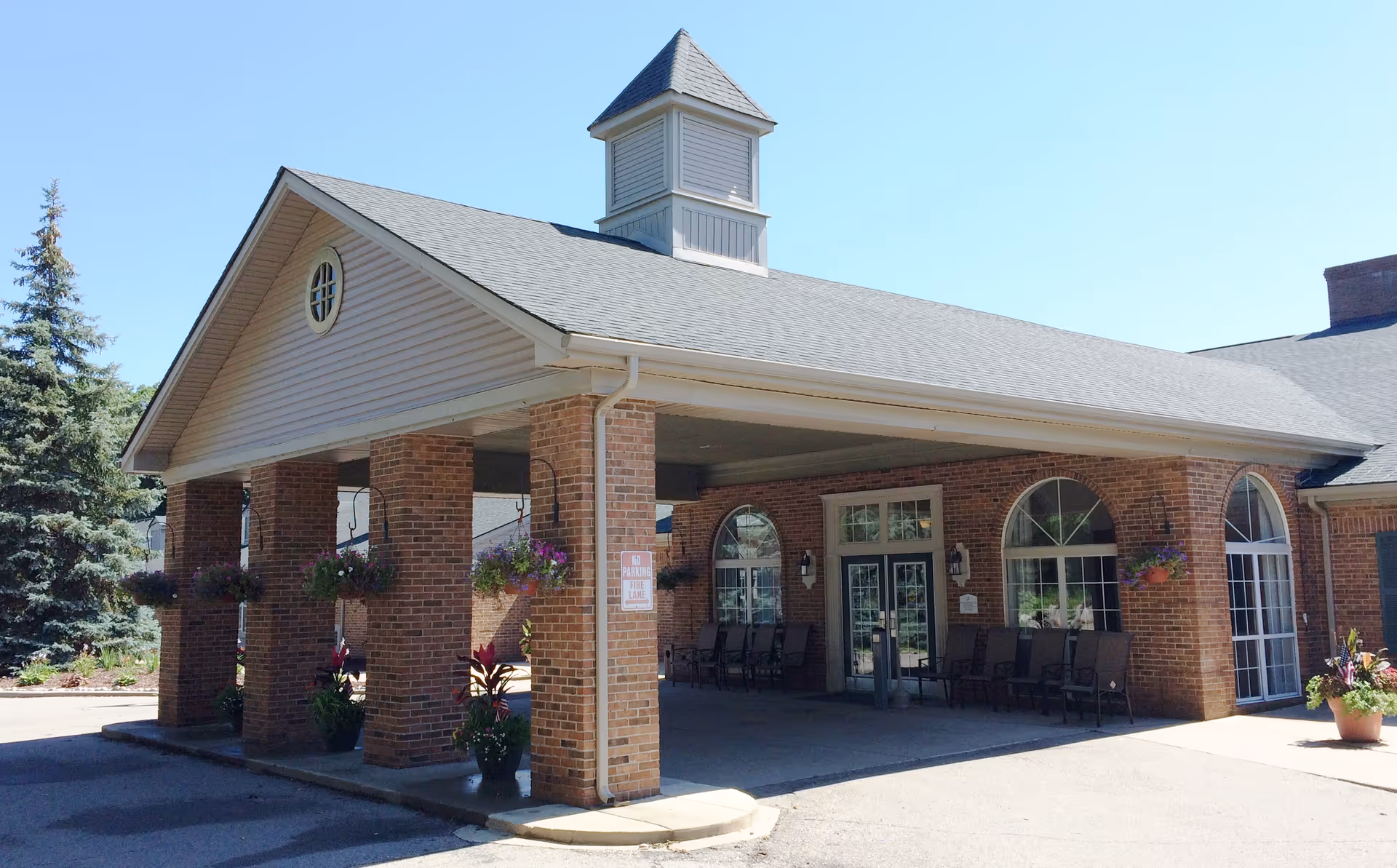 Exterior view of a brick building with a covered entrance supported by brick columns. The building has large arched windows and a small cupola on the roof. There are hanging flower baskets and potted plants around the entrance area.