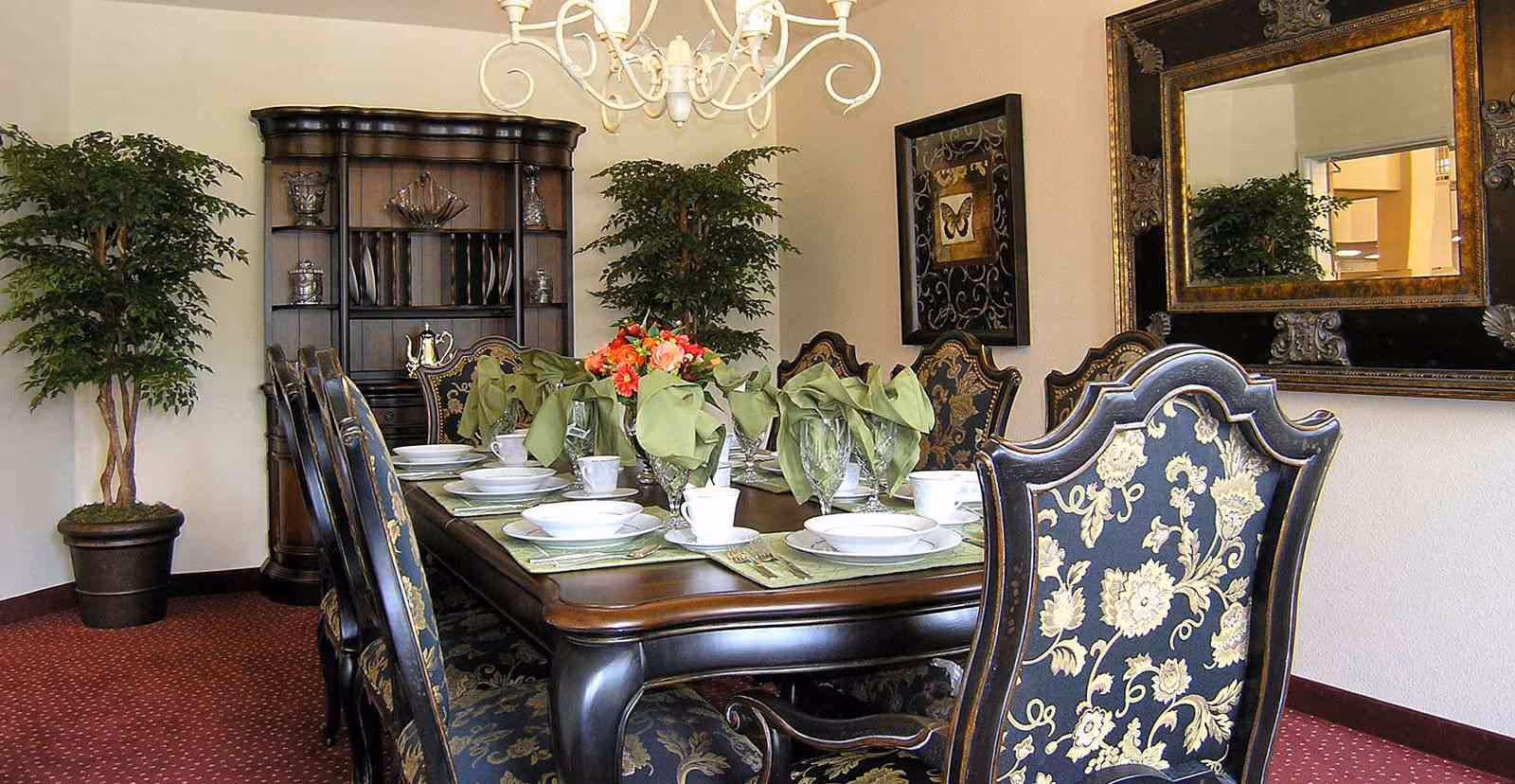 Elegant dining room with a dark wooden table set for eight, featuring white plates, cups, crystal glasses, and green cloth napkins. The chairs have ornate floral upholstery. The room includes a large decorative mirror, framed butterfly artwork, two potted plants, a dark wooden cabinet with glassware, and a white chandelier overhead.