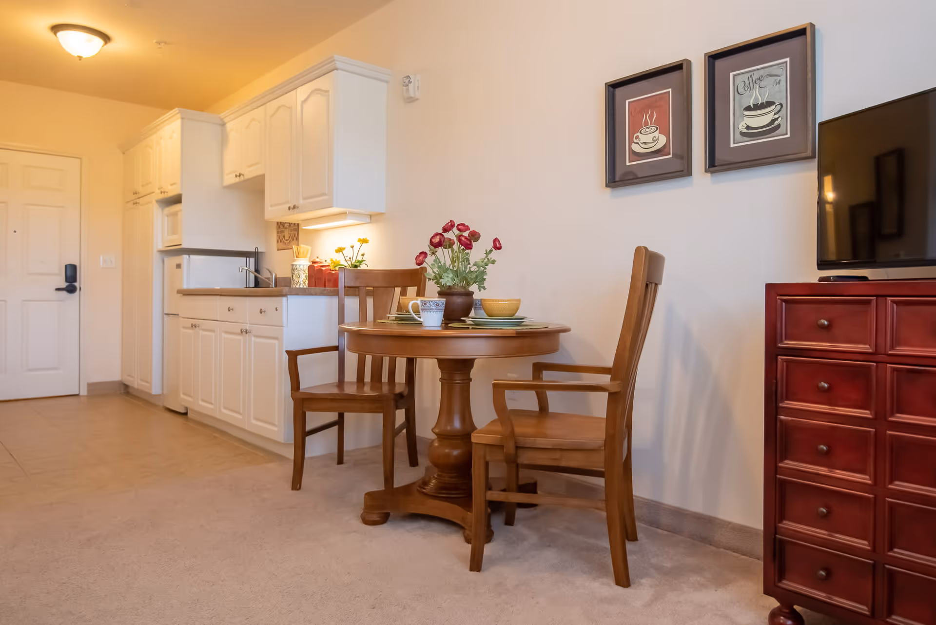 A small dining area in a senior living facility with a round wooden table set for two, featuring a flower pot with red flowers, cups, and bowls. The area includes two wooden chairs, a red chest of drawers with a flat-screen TV on top, and a kitchenette with white cabinets, a microwave, and a refrigerator. The walls are decorated with two framed coffee-themed pictures.