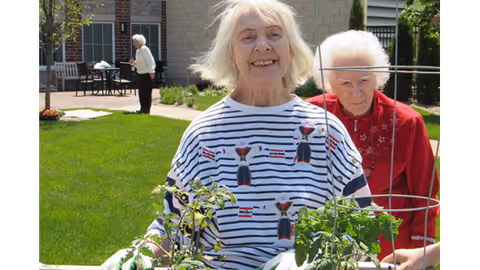 Two elderly women gardening outdoors in a green lawn area with plants and gardening tools. One woman in a striped shirt is smiling at the camera, while the other woman in a red jacket is focused on the plants. In the background, another person is walking near a building with large windows and patio furniture.