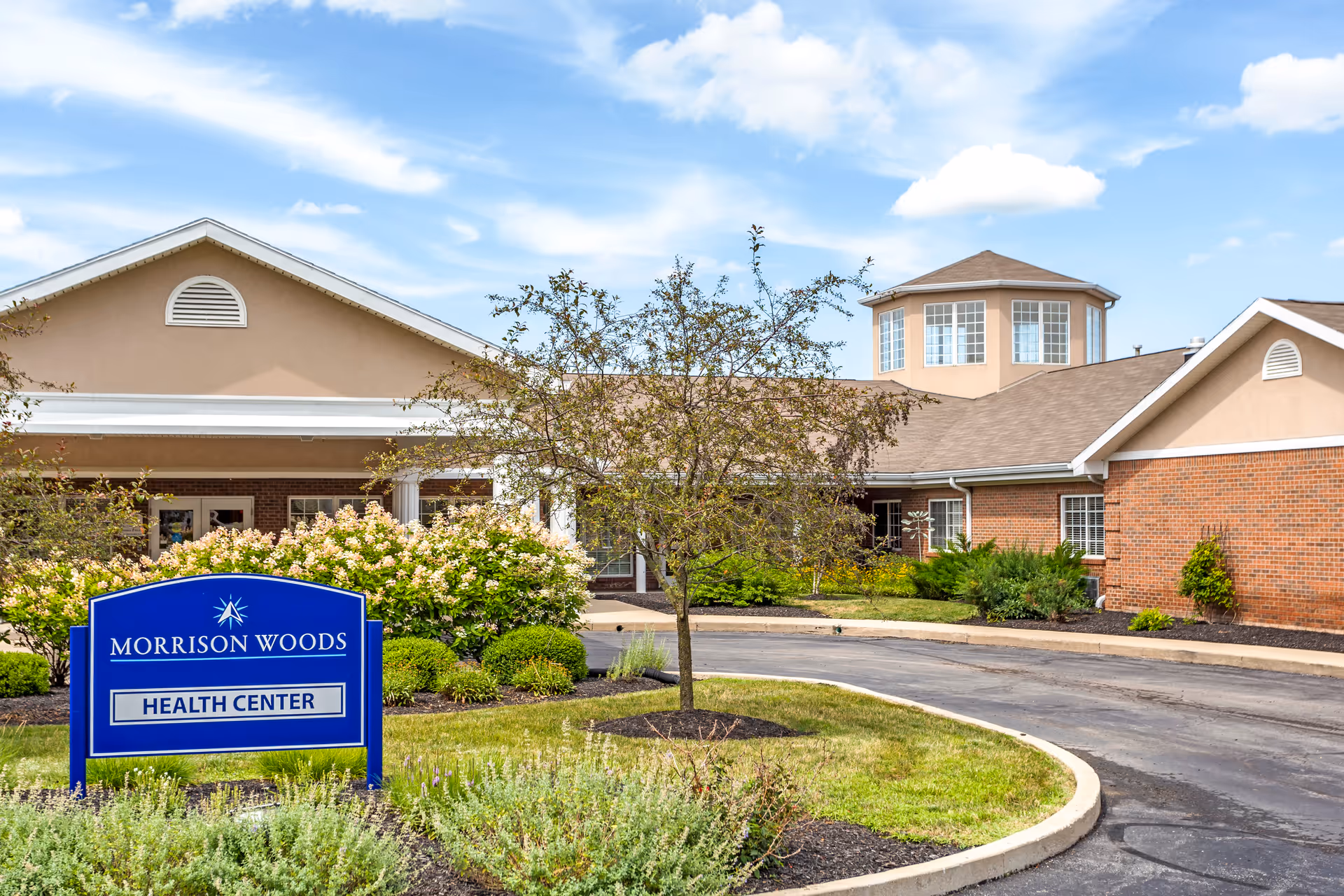 Exterior view of Morrison Woods Health Center building with a blue sign in front surrounded by greenery and a curved driveway. The building has a beige and brick facade with a small tree and bushes in the landscaped area.