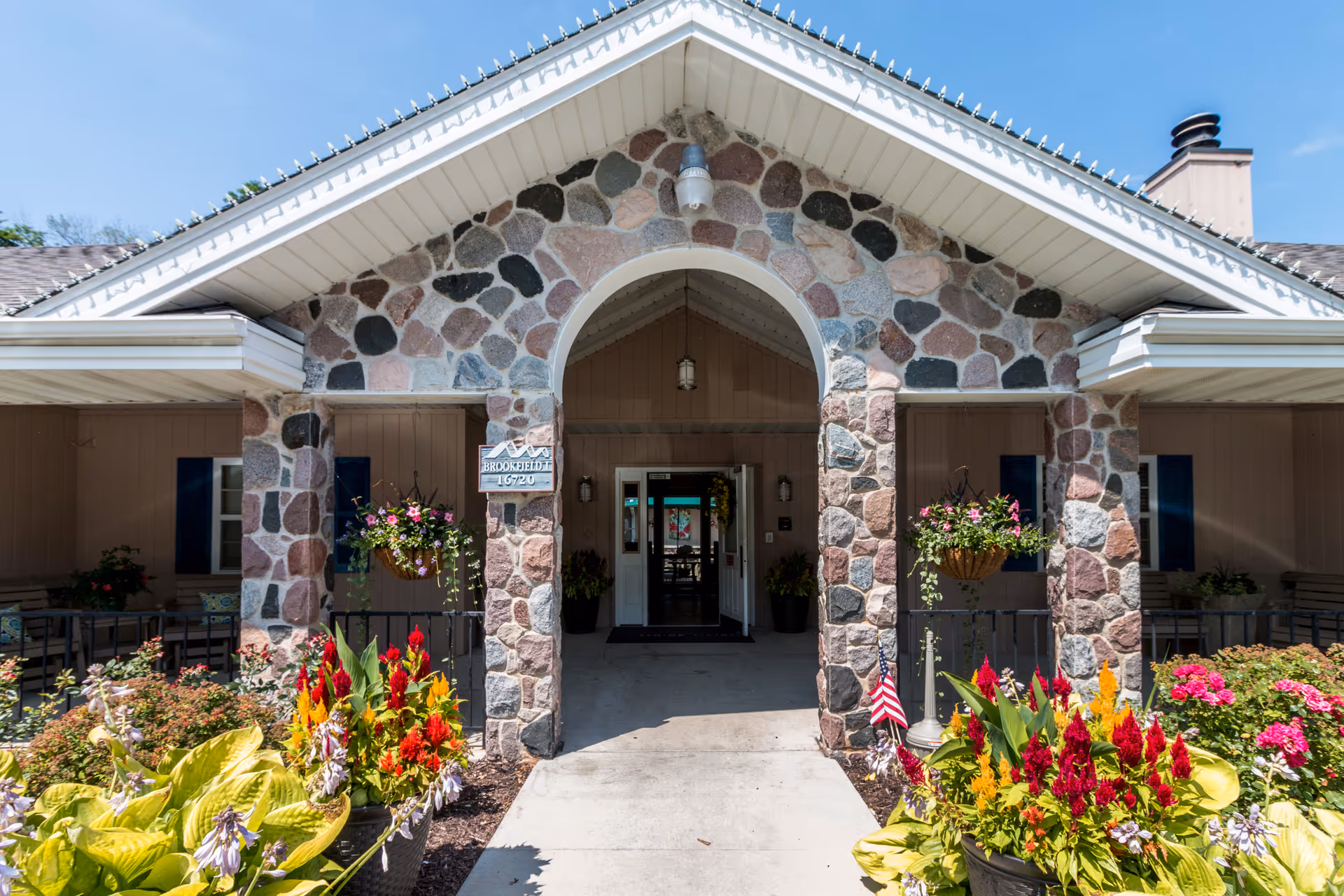 Entrance to a senior living facility with a stone archway and pillars, colorful flower beds on either side of the walkway, and a clear blue sky above.