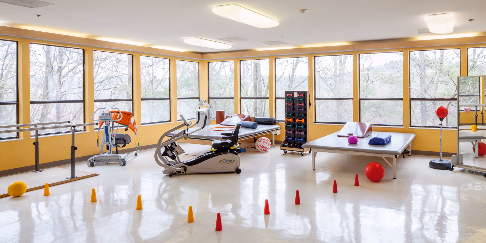 A bright physical therapy or rehabilitation room with large windows showing trees outside. The room contains exercise equipment including a stationary bike, parallel bars, therapy tables with towels and exercise balls, a rack of colorful dumbbells, and small orange and red cones arranged on the floor.