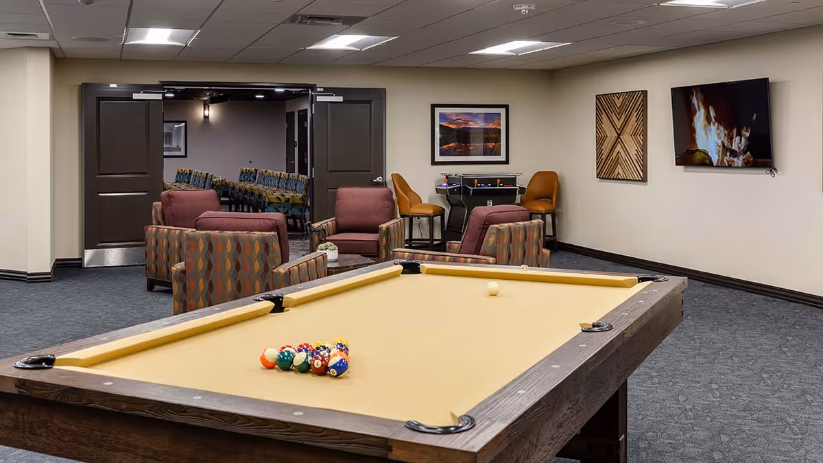 A senior living facility game room featuring a pool table with balls set up for a game. Behind the pool table are several comfortable armchairs and a small table with a plant. On the wall, there is a flat-screen TV displaying a fireplace scene, a framed landscape photograph, and a decorative wooden art piece. In the background, double doors open to another room with patterned chairs arranged in rows.